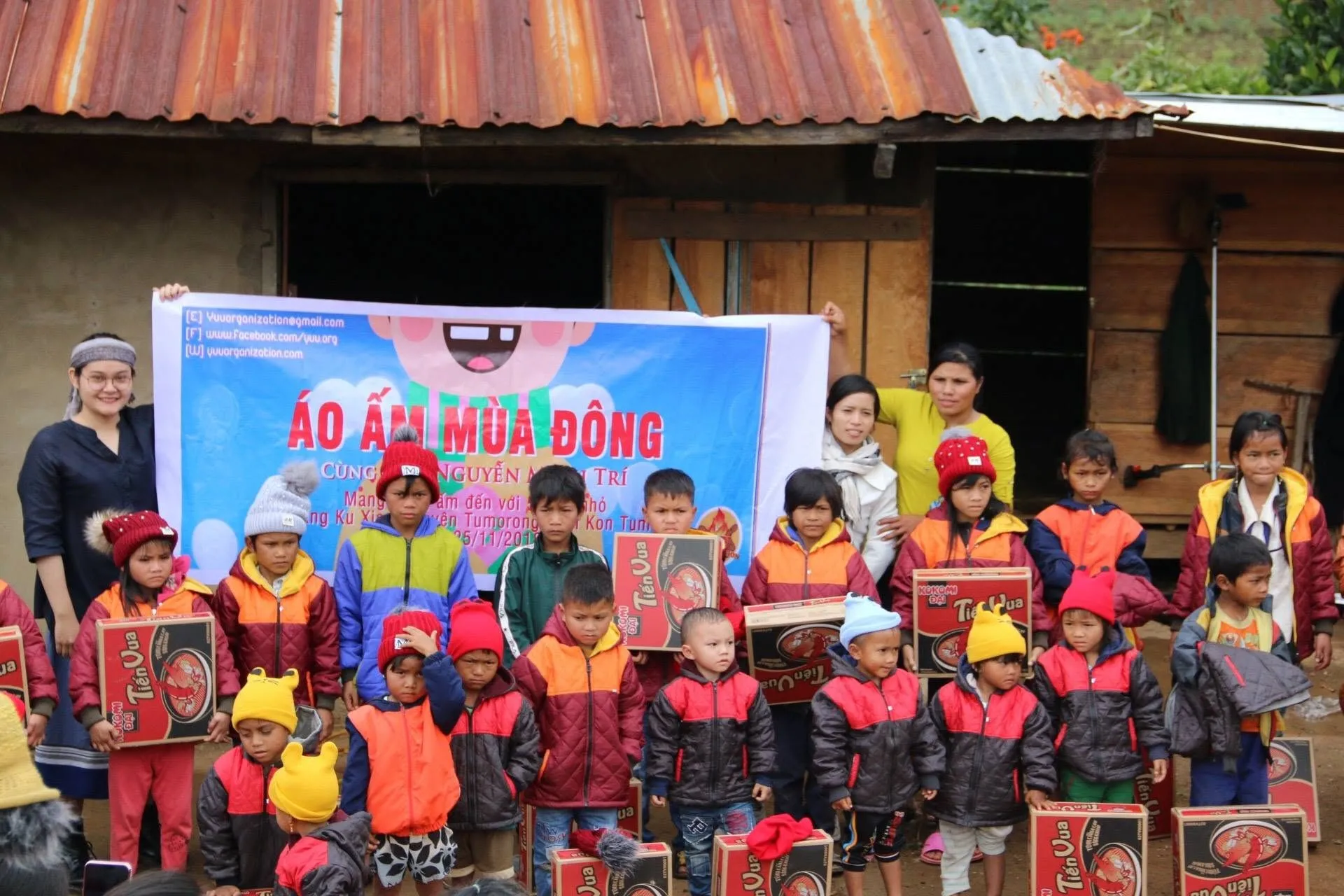 Children in new warm jackets and hats holding gift boxes in front of the Áo Ấm Mùa Đông banner