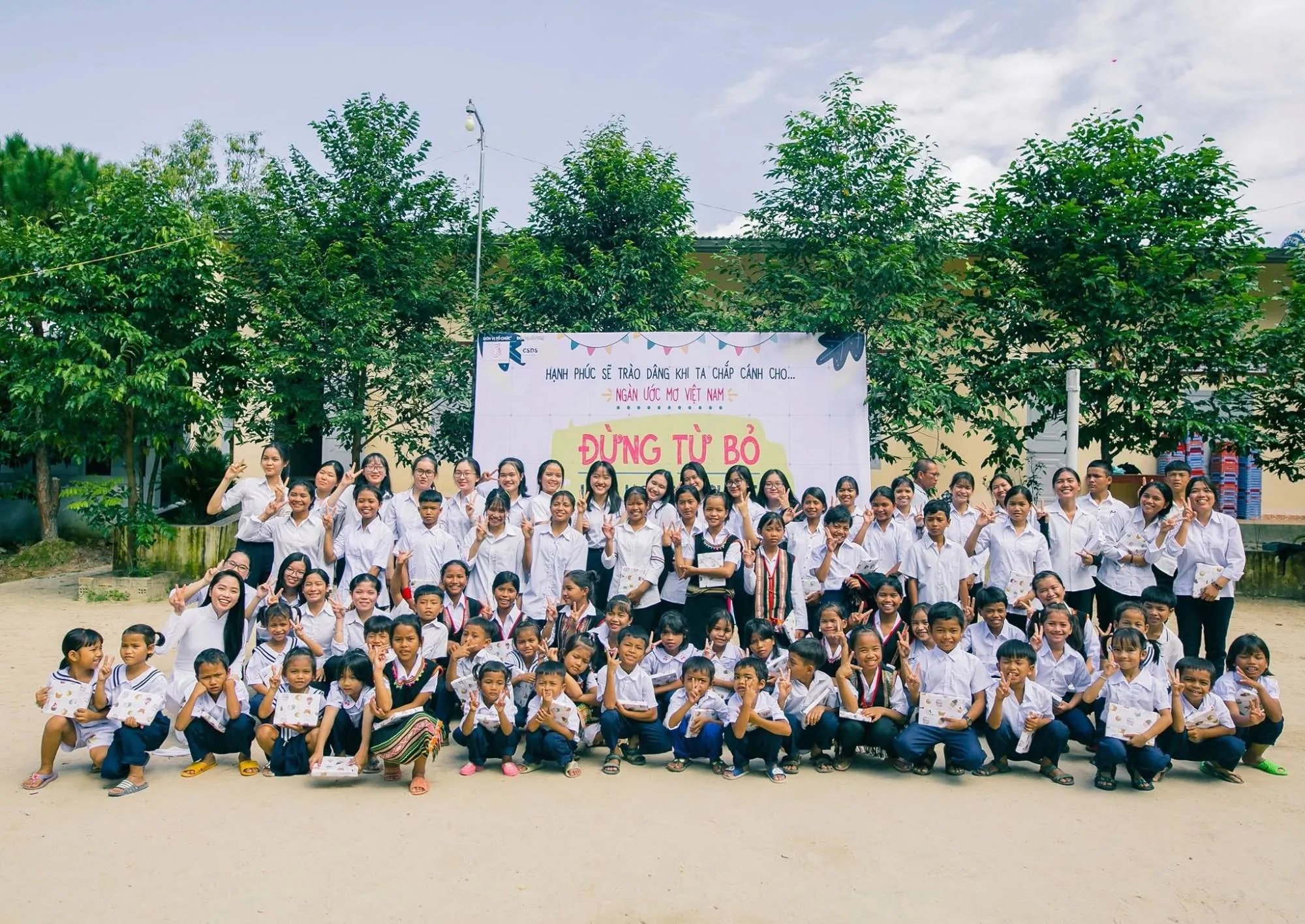 Students in white shirts and volunteers making the V sign in front of the Đừng Từ Bỏ banner