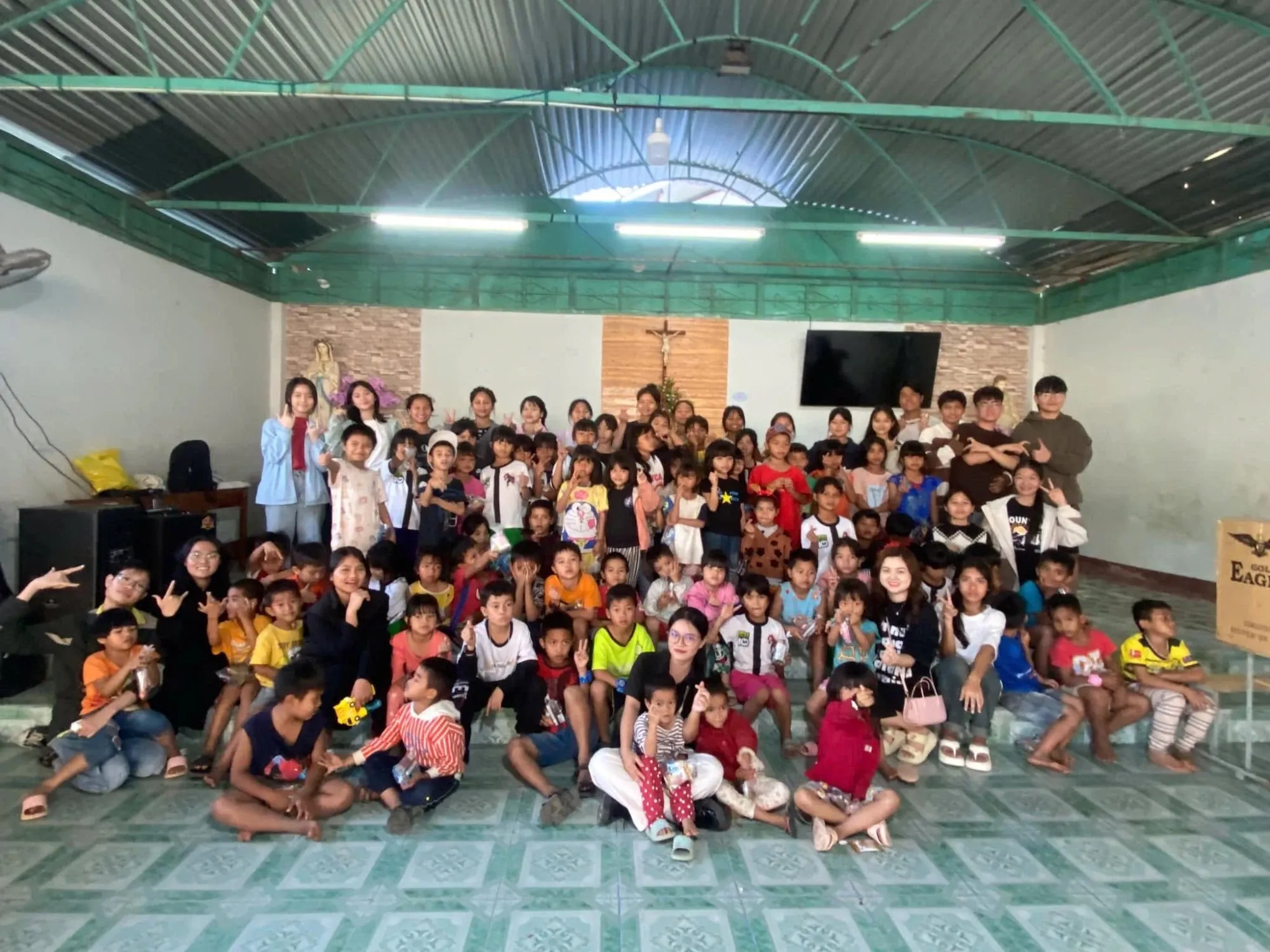 Volunteers and children of Vinh Sơn 2 together in a hall, waving to the camera